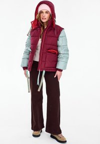 Nő bordó és szürke kapucnis steppelt dzsekiben, rózsaszín sapkában, krémszínű pulóverben, sötét nadrágban és világosbarna túracipőben, fehér háttér előtt áll.