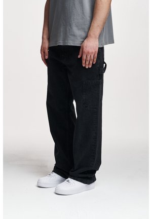 Homme portant un pantalon cargo noir et des baskets blanches, debout avec les mains détendues le long du corps devant un fond uni.