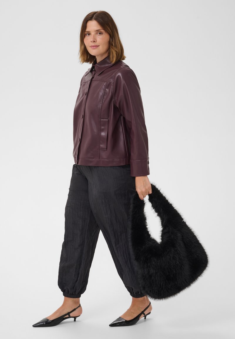 Femme portant une veste en cuir violet foncé et un pantalon noir texturé, tenant un grand sac à main noir duveteux, debout devant un fond uni.