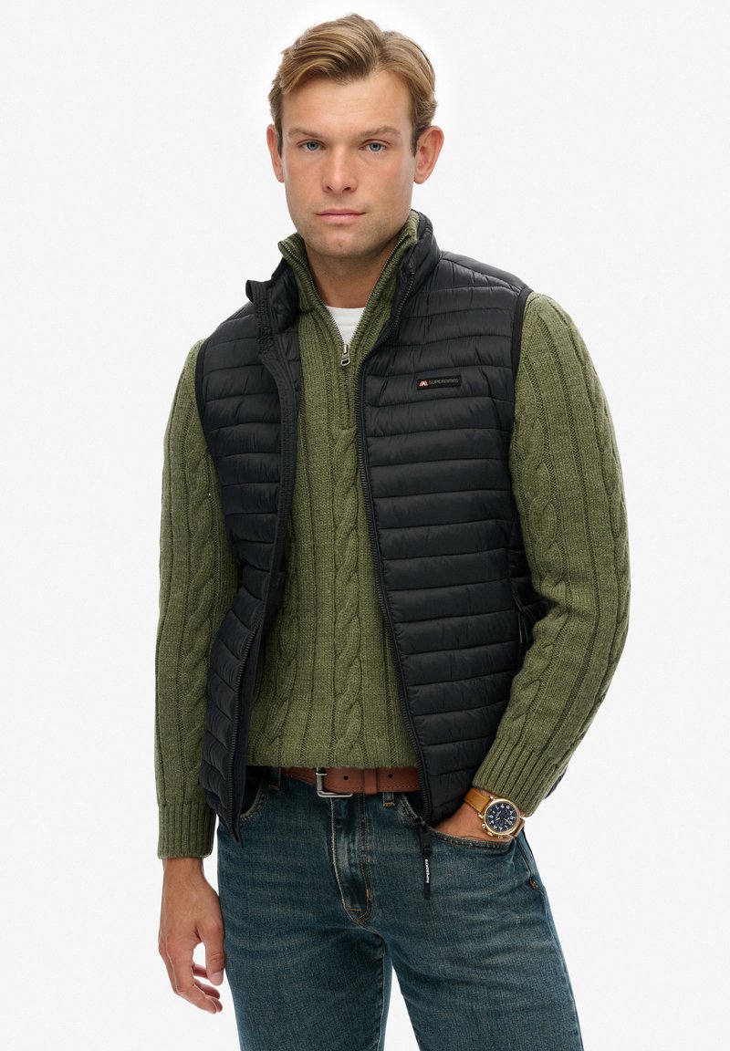 Chaleco acolchado negro sobre un suéter de punto en verde. El chaleco presenta un cierre frontal con cremallera y un diseño ajustado. Jeans de mezclilla azul y un cinturón marrón.