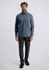 Un uomo in piedi indossa un pullover blu a maniche lunghe con zip, pantaloni grigio scuro e scarpe di pelle nere su uno sfondo chiaro e semplice.