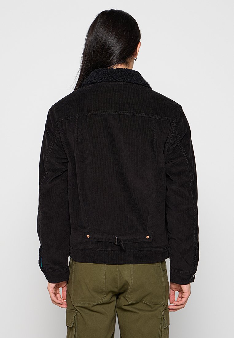 Chaqueta de pana negra con cuello de borrego, acentos de botones y textura acanalada. Presenta un diseño ajustado y una correa ajustable en la parte posterior.