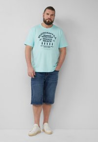 Mintgroene t-shirt met marineblauwe bedrukking met de tekst “VENICE BEACH,” gecombineerd met knie-lange denim shorts en witte sneakers. Effen achtergrond.