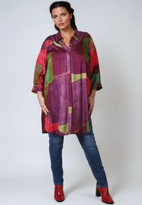 Frau mit dunklem Haar, die ein lockeres, mehrfarbiges Kleid in Lila, Rot und Grün trägt, dazu Jeans und rote High Heels. Sie steht vor einem einfarbigen Hintergrund.