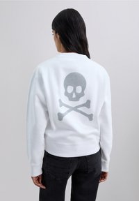 Woman with long dark hair wearing white sweatshirt with gray skull and crossbones graphic on the back, paired with black jeans.