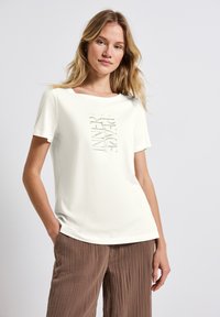 T-shirt crème clair en coton doux, avec un imprimé graphique centré dans un ton atténué. Le design a une coupe décontractée et un col large.
