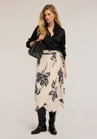Femeie blondă îmbrăcată cu o bluză din satin negru, o fustă înflorată bej cu model și cizme negre până la genunchi, ținând o geantă neagră pe un fundal neutru.