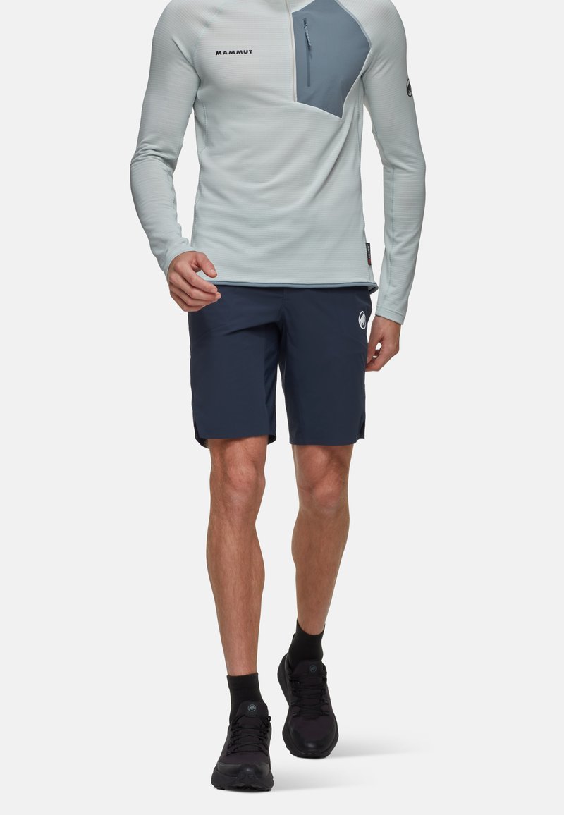 Ljusblå långärmad tröja med en grå fickdetalj, parat med marinblå shorts med en logotyp, bärs med svarta skor och strumpor.