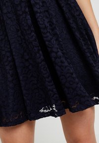 Jupe en dentelle bleu foncé à motif floral portée au-dessus des genoux, montrant une partie des jambes et un bras sur un fond uni.