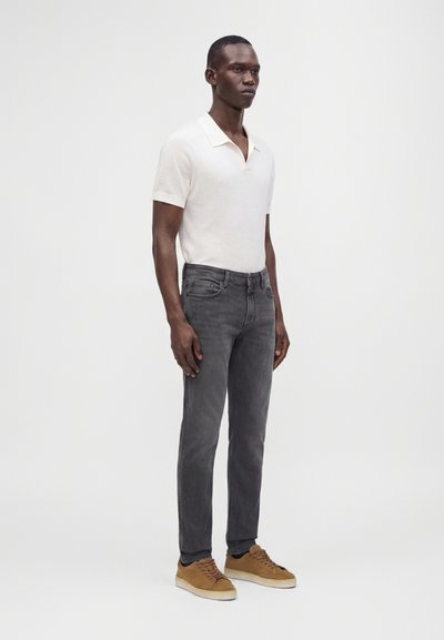 Homme debout portant un polo blanc à manches courtes, un jean slim gris et des baskets marron sur un fond uni et clair.