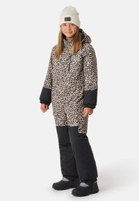 Schneeanzug mit Leopardenmuster, beige-schwarzes Design, mit elastischen Bündchen, Reißverschluss vorne und flachen Taschen. Kombiniert mit einer weißen Strickmütze und schwarzen Stiefeln.