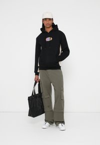 Sudadera negra con logo multicolor, paneles laterales beige, pantalones cargo oliva con bolsillos, mochila negra, zapatillas blancas, gorra de color claro.
