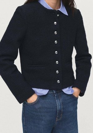 Kvinde iført en sort tekstureret jakke med sølvknapper over en blå stribet skjorte, hænder i lommerne, kombineret med blå jeans.