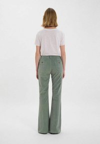 Pantalons en velours côtelé vert évasés avec une finition texturée, featuring deux poches arrière et des détails de boutons, associés à un t-shirt de couleur claire.