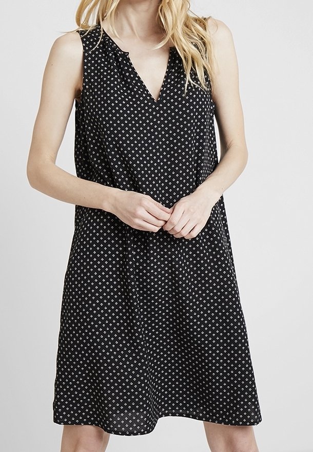 Robe noire sans manches avec motif à losanges blancs, coupe ample, portée par une personne aux cheveux blonds, les mains croisées devant.