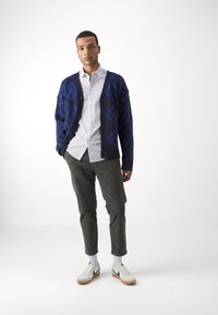 Marineblauer, gemusterter Strickcardigan über einem weißen Hemd mit Knöpfen, kombiniert mit olivgrünen Cordhosen und weißen Sneakern.