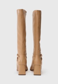 Bottes en simili cuir beiges jusqu'aux genoux avec des talons carrés, dotées de deux boucles latérales et d'une fermeture éclair à l'arrière pour un enfilage facile.