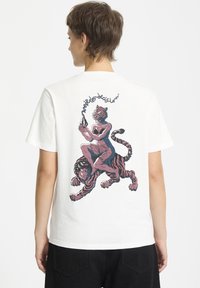 Hvid bomulds T-shirt med et farverigt grafik på ryggen, der viser en figur med en tigerhoved, siddende på en tiger, med kraftige konturer.