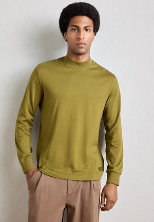 T-shirt en tricot à manches longues, vert olive, avec un col haut et des poignets côtelés. Présente une texture subtile et une étiquette sur l'ourlet.