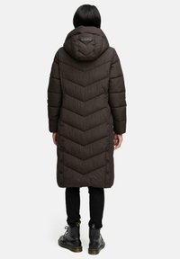 Lungo cappotto imbottito marrone con un motivo a zig zag, cappuccio e bottoni neri lungo il lato, indossato con leggings neri e stivali.