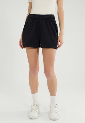 Eine Person trägt schwarze elastische Shorts mit elastischem Bund, weiße Socken, weiße Turnschuhe und ein weißes kurzärmliges Oberteil vor einem hellen Hintergrund.