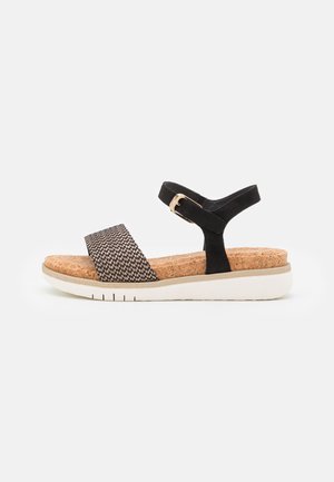 Sort og beige sandal med åben tå med flettet frontrem, korksål, ankelrem med guldlås og hvid rillede sål.