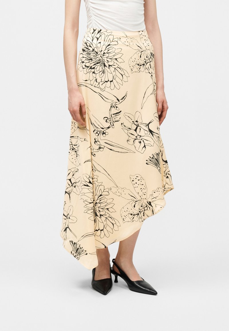 Μπεζ ασύμμετρη midi φούστα με μαύρο σκίτσο λουλουδιών, συνδυασμένη με μαύρα πέδιλα slingback και λευκό αμάνικο τοπ.