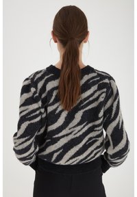 Kvinna med lång brun hästsvans som bär en svart och grå randig fluffig tröja, vänd bort mot en neutral bakgrund.