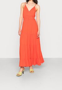 Robe maxi de couleur corail vif, avec un décolleté en V, des bretelles ajustables, une taille froncée et un ourlet à volants en plusieurs niveaux. Associée à des sandales à talons blocs vertes.
