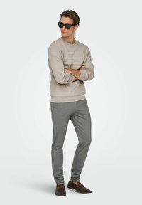 Pull beige texturé, pantalon gris ajusté, et lunettes de soleil foncées. Des mocassins marron avec des accents métalliques dorés complètent le look.