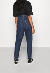 Modré džíny s vysokým pasem a elastickým pasem, zadní kapsy s červenou značkou loga, v kombinaci s černými teniskami.