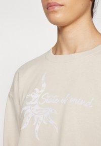 Beige sweatshirt med ribbad rund halsringning, prydd med en vit solgrafik och texten "State of mind" i en avslappnad stil.
