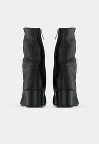 Ζευγάρι μαύρα δερμάτινα μποτάκια αστραγάλου με τακούνια τύπου block και φερμουάρ στο πίσω μέρος, εμφανιζόμενα από πίσω σε λευκό φόντο.