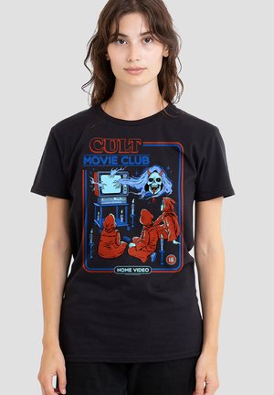 Person wearing a black shirt with a graphic of three red-hooded figures watching a vintage TV showing a ghostly skull image.