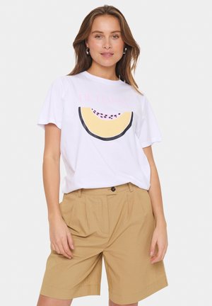 Camiseta blanca con un gran gráfico de sandía amarilla y el texto "SE FELIZ". Pantalones cortos beige con pliegues. Modelo de pie contra un fondo neutro.