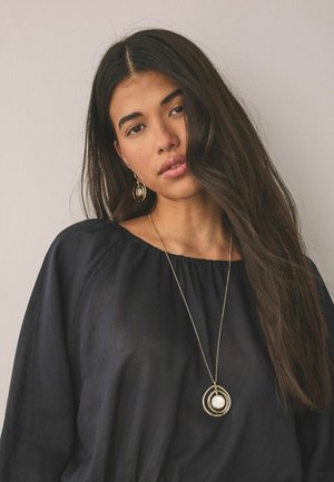 Young woman with long dark hair wears black blouse, large gold hoop earrings, and gold pendant necklace with circular design and white center.