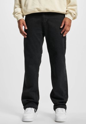 Personne portant un jean noir ample, des baskets blanches et un haut beige à manches longues, debout devant un fond uni.
