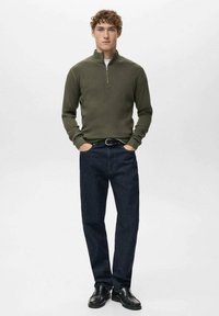 Jersey de punto acanalado color verde oliva con cuello de cremallera hasta un cuarto, combinado con vaqueros oscuros y zapatos negros. Silueta sencilla y entallada.