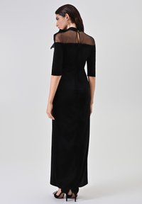 Zwarte avondjurk met een transparant rugpaneel, ellebooglange mouwen, een lange silhouet en een getailleerde taille. Voorzien van een knoopsluiting bij de hals.