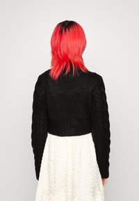 Osoba z jasnoczerwonymi włosami, ubrana w czarny, dziany crop top i fakturowaną białą spódnicę, odwrócona plecami na tle jasnym.