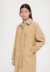 Ung kvinna med rakt svart hår iklädd en beige knappad kappa över en grå polotröja, stående med en hand i fickan.
