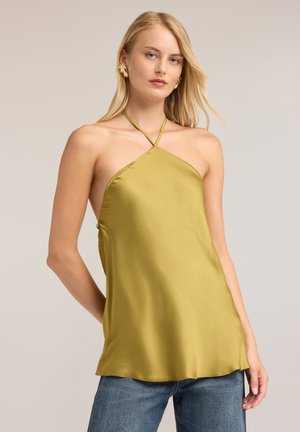 Femme blonde portant un haut dos nu jaune moutarde avec un décolleté à fines bretelles et un jean bleu sur un fond neutre.