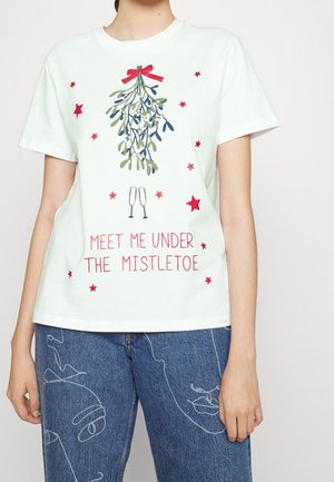 Biały t-shirt z grafiką jemioły i czerwonych gwiazd, dwie szampanówki i czerwony tekst z napisem "Meet me under the mistletoe", noszony z niebieskimi jeansami.