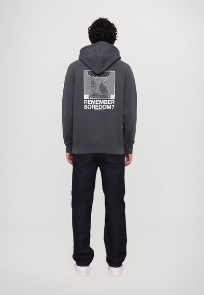 Grijze hoodie met een voorzak, voorzien van een grafische afdruk op de rug met de tekst "VERGEET JE BOREN?" in combinatie met donkere jeans en witte schoenen.