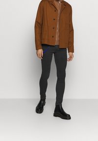 Man som bär en brun jacka över en brun dragkedjetröja, svarta smala byxor och svarta läderstövlar, stående mot en enkel ljus bakgrund.