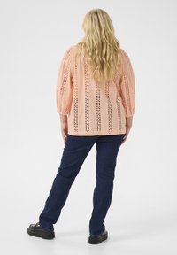 Peach-colored blouse with intricate eyelet design, three-quarter sleeves, paired with dark blue jeans and black shoes.