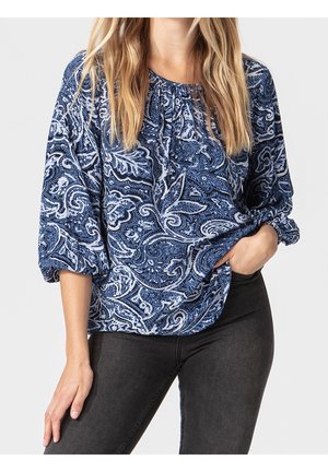 Nainen pukeutuneena siniseen paisley-kuvioiseen kolmivartiseen paitaan, yhdistettynä mustiin istuviin farkkuihin.