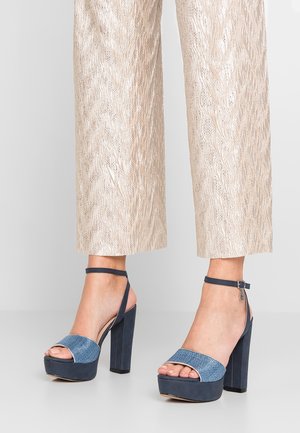 Stopy w ciemnoniebieskich sandałach na platformie na wysokim obcasie z paskami na kostkach, w połączeniu z kremowymi szerokimi spodniami o fakturze.