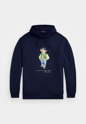 Marineblauwe hoodie met een beer die een groene varsityjas, spijkerbroek, sneakers en een blauwe pet achterstevoren draagt, met de tekst Polo Bear by Ralph Lauren eronder.