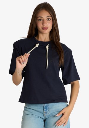 Giovane donna con lunghi capelli castani che indossa una maglietta blu navy a maniche corte con cordoncini bianchi e jeans azzurri su sfondo bianco.